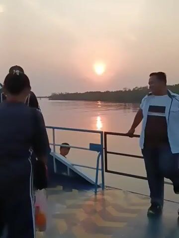 The video shows a group of people on a boat during sunset. The sun is setting over a calm body of water, casting a warm glow on the scene. Several individuals are visible, some standing and looking out at the water, while others are in conversation. One person is leaning on a railing, and another is holding a bag. The boat appears to be a ferry or similar passenger vessel, with visible seating and railings. The overall atmosphere seems relaxed and peaceful as people enjoy the natural beauty of the sunset.