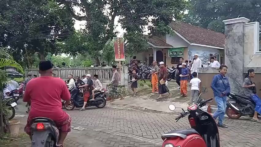 The video shows a scene of people gathering outside a building in a residential area. The majority of the people are men and boys, many wearing white caps, and some are dressed in traditional clothing. Several motorcycles are parked along the paved road, and people are either walking or riding on them. The building appears to be a house or a small community center, and there are trees providing shade in the background. The atmosphere seems lively and community-oriented.