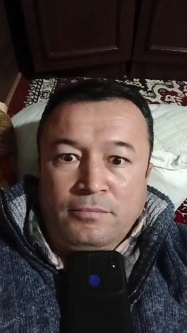 A man is lying down looking directly at the camera. He is holding a black smartphone with a blue notification light. He has dark hair and brown eyes, and is wearing a blue hooded sweatshirt. He is lying on a white pillow. A red patterned rug is visible in the background. Above him are dark wooden cabinets.