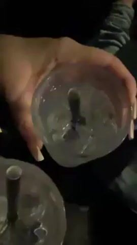 The video shows a group of people at an indoor gathering, likely a bar or party. Two clear plastic cups filled with liquid and ice, each with a black straw, are held in someone's hands in the foreground. The camera pans up to show a group of people raising their drinks in a toast. Individuals are wearing casual clothing. The lighting is dim.