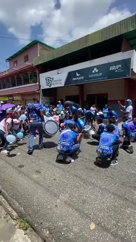The video shows a group of people playing drums in the street. They are dressed in blue shirts with a logo on the back. The group is surrounded by people, some holding blue and white balloons. They are on a street, in front of a store. The video seems to be taken during a community celebration.