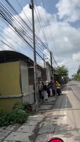 A group of people, mostly women and a few children, are gathered on a sidewalk next to a street. Some are standing near a building with a sign that reads "DEWAN QURBAN." A woman on a motorbike is also present, facing the group. The scene appears to be outdoors, with power lines overhead and a cloudy sky.