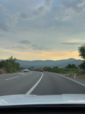 This media file shows a view from inside a vehicle traveling on a highway in Liješnje, Montenegro. The highway curves to the right, with lush green trees and bushes lining the sides. In the distance, rolling hills and mountains are visible, some appearing forested. The sky is partly cloudy with a blend of soft blues and warm yellows near the horizon, suggesting it is either sunrise or sunset.

Several cars are driving on the highway ahead. A white car is in the left lane, followed by a black car, a dark sedan, and a red car. A large billboard is positioned on the right side of the road, displaying an image of a bird and text that reads "BIOMEĐO WILDLIFE PARK". Below this, there is smaller text that is difficult to read, but appears to be an advertisement.

The overall scene conveys a sense of travel and scenic beauty. The time of day appears to be late afternoon or early evening, given the warm glow in the sky. The weather seems pleasant, with dramatic clouds adding to the visual appeal of the landscape.