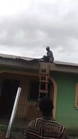 The video shows a man sitting on a roof, near the edge of a building with a green exterior wall and a metal roof. He is wearing a dark shirt, dark pants, and a green cap. A wooden ladder leans against the building, leading up to where the man is sitting. There is a metal pole propped up on the ground near the building. Another man, partially visible in the foreground, is wearing a patterned shirt. The sky is overcast. The man on the roof appears to be working on the roof, arranging pieces of metal, possibly roofing sheets. The person filming the video is standing on the ground, recording the scene.