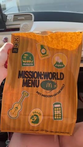 A person is holding a McDonald's paper bag, which is orange with brown stripes. The bag has the text "MISSION: WORLD MENU" and "MIT MECCES UM DIE WELT" printed on it. It also features stickers representing different countries and items: Malaysia, Canada, Japan, a guitar, Brazil, and what appears to be a calculator. The bag is inside a car, with the dashboard visible in the background.