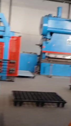 This is a video showing a warehouse or factory setting. In the foreground, there are stacks of large, flat, metallic sheets, possibly aluminum or steel. These sheets are neatly arranged on shelves and on a table-like surface. The sheets have a clean, reflective surface. In the middle ground, there's industrial machinery. One machine is prominent, painted blue with a large red metal grid structure in front of it. Another machine, gray in color, is also visible with some yellow warning labels. Behind these machines, more shelves are visible, stacked with similar metallic sheets, some of which appear to be wrapped in plastic. The environment is lit by overhead lights, typical of an industrial space, and the floor is concrete. There are no people visible in the frame. The video appears to be a pan across the workspace, focusing on the material and the equipment used for processing or storing it.
