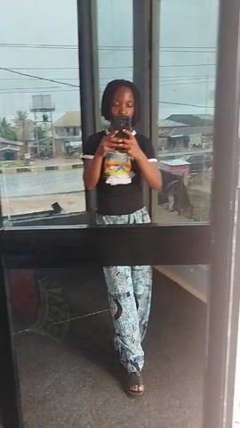 The video shows a young person standing in front of a glass door and taking a selfie with their phone. The person is wearing a black t-shirt with a colorful graphic on it and patterned pants. Their reflection is visible in the glass, showing them from the waist up, and their full body is visible in the reflection on the floor. Outside the glass, a street scene is visible, with buildings, cars, and a cloudy sky. The person moves slightly, shifting their weight from one foot to the other.