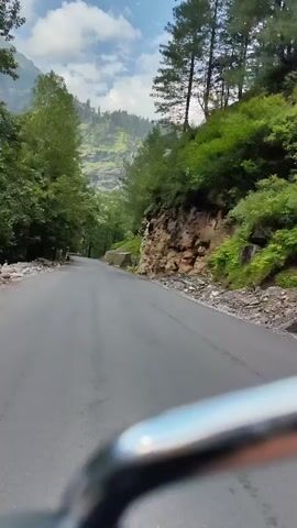 A first-person perspective video shows a paved road winding through a dense forest with lush green trees on either side. The road appears to be a mountain road, with steep, rocky slopes and dense vegetation on the right side. The sky is partly cloudy, with patches of blue visible. The video appears to be shot from a moving vehicle, possibly a motorcycle or bicycle, with the handlebars visible in the foreground. The road surface is wet in some areas, suggesting recent rain. The overall impression is one of a scenic drive through a natural landscape.