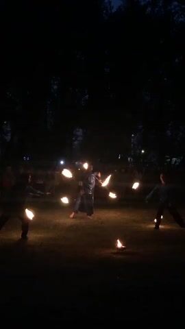 This video captures a nighttime outdoor performance featuring three individuals engaged in fire juggling. The scene is dark, with the primary light source emanating from the performers' props. Two individuals are positioned on the left and right sides of the frame, while a third performer is centered and slightly further back, wearing a dark robe or cloak. All three are actively manipulating flaming objects, creating arcs and trails of light. The ground appears to be grass or dirt, and in the background, silhouettes of trees are visible against a dark sky. There are also faint lights in the background, possibly from spectators or distant structures. The performance seems to be taking place in a park or open field.