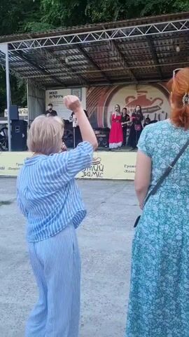 A medium shot captures an outdoor scene where a band performs on a stage while two women dance in the foreground, their backs mostly to the camera. The band, consisting of at least five members, plays instruments like an accordion and guitar, with one female vocalist in a red dress standing prominently. A banner with "МОЇ ПОРУЧ" and a logo featuring a stylized pot is visible behind the band. The dancing women, both with light-colored hair, move to the music. One woman, in a blue striped shirt and striped pants, raises her arm and sways. The other woman, in a dark embroidered top and gray pants, dances facing the first woman. To the right, another woman in a floral dress stands watching. The setting appears to be a park or outdoor event space with trees visible in the background.
