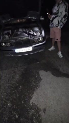 The video shows two young men outdoors at night. One is standing next to a silver car with its hood open, looking at his phone. The other is walking toward a black SUV parked nearby, also looking at his phone. The ground appears to be gravel. There's a full moon visible in the sky.