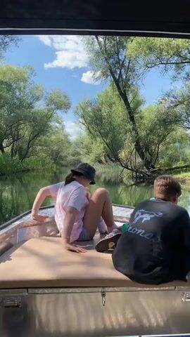 Three individuals are on a boat, traveling down a calm river surrounded by lush green trees and foliage. The scene appears to be set during the daytime, under a bright sky with scattered clouds, suggesting pleasant weather. The boat, made of metal with a padded seating area, has a black canopy providing shade.

The subjects are positioned towards the back of the boat. On the left, a person wearing a pink t-shirt and a black bucket hat is seen from the side. In the center, a young person in a blue t-shirt is also facing forward. On the right, another young person in a black t-shirt with a graphic design on the back is seated, holding what appears to be a fishing rod.

The activity taking place seems to be a leisurely boat ride, possibly combined with fishing, given the presence of the fishing rod. The river is tranquil, reflecting the surrounding trees and sky. The vegetation is dense, indicating a natural, possibly somewhat wild, environment. The overall mood is relaxed and peaceful.

On the back of the black t-shirt, a graphic of a horse or bull is visible, along with some text below it, which is not clearly legible. The time of day is suggested by the bright sunlight and the clear sky. The location context of Cerovi Pod, Montenegro, points to a natural landscape characteristic of the region.