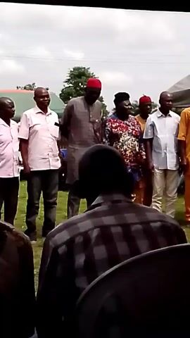 This video shows a group of men and one woman standing outside. Most of the men are wearing light-colored shirts and dark pants. One man in the center is wearing a patterned tunic and a red hat. The woman is wearing a colorful patterned dress. The group appears to be facing a crowd, and some individuals in the foreground are obscured by their backs. The setting is outdoors, with grass and trees visible in the background. There are also tents or shelters in the distance. The lighting suggests it is daytime.