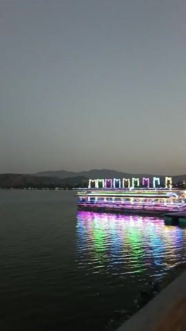 The video displays a panoramic view of a large body of water, likely a lake, under a dusky, overcast sky. Two illuminated boats are visible on the water, moving from left to right across the frame. The boat on the left is closer to the viewer and is decorated with colorful, bright lights reflecting on the water. The boat on the right is further away and also brightly lit with a sequence of multicolored lights. In the distance, a treeline forms the horizon, with faint outlines of hills or mountains beyond. The water surface is calm with gentle ripples. The lighting suggests it is either dusk or early evening.