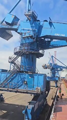 A large blue crane with the markings "ABP Siwertell CSU 144" is seen from the side, angled upwards. Below it, a ship is docked, its cargo hold filled with light-colored bulk material. The crane appears to be in the process of unloading or loading, as a large, rectangular, grey component is being lowered into the ship's hold. In the background, other cranes and port equipment are visible under a bright, partly cloudy sky. The scene takes place at a port, with evidence of industrial activity. There are no visible people interacting directly with the crane or the ship at the moment captured in the video.