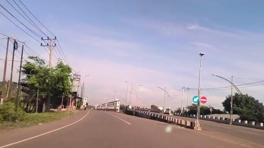 A car drives down a road toward a metal bridge under a blue sky with wispy clouds. The road is paved and has white lines marking the lanes. On the left side of the road is a building with a dark gray roof and a wooden structure. Power lines and poles run along the left side of the road. On the right side of the road is a low wall topped with white concrete blocks, and there are streetlights with arched tops. There is a sign showing a blue arrow pointing left and a red circle with a horizontal line in it. As the car approaches the bridge, a motorcyclist in a white shirt can be seen crossing the bridge. The bridge itself is made of white metal and has a triangular pattern in its structure. There is another bridge visible behind the first one.