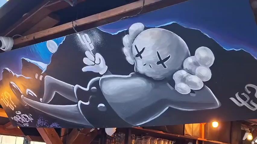 The video opens with a close-up of a large mural painted on a wooden surface, depicting a stylized grey cartoon character with X's for eyes. The mural appears to be hanging from a wooden structure, possibly a ceiling or overhang. The character is depicted in a relaxed, almost lounging pose, with one hand extended upwards. The background of the mural is a dark, abstract landscape in shades of blue and purple.

The camera then pans to the right and shows a blurry outdoor scene. There are lights visible, possibly string lights, and some greenery. The scene is out of focus and quickly transitions.

The video then focuses on a large, black, balloon-like sculpture of a character with X's for eyes, similar to the one in the mural. This sculpture is decorated with various graffiti tags and drawings in bright colors, including words like "OK," "POP," and "WHITE." Several people are gathered around the sculpture, some of whom are looking at it or taking pictures. The environment appears to be an outdoor public space, with wooden seating and some trees visible in the background. The lighting suggests it is daytime or early evening. One person in the foreground is raising their hands, and a child is visible in the lower right corner. The overall atmosphere seems casual and social.
