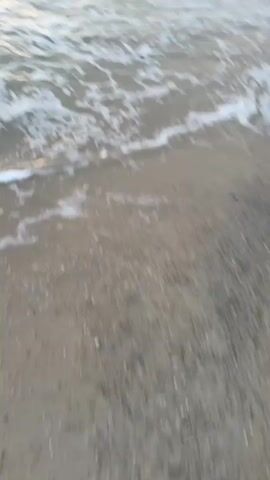 This video captures a sunset at a beach. The camera is positioned low to the ground, looking out towards the ocean. The waves are breaking gently on the sandy shore. The sky is filled with dramatic clouds, illuminated by the warm glow of the setting sun. The colors in the sky range from soft oranges and yellows to deeper grays. The water reflects the colors of the sunset, creating a beautiful and serene scene.