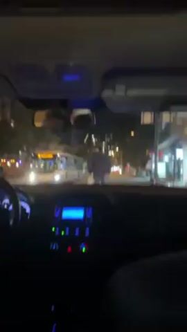 From within a car, the video shows a man wearing a grey hoodie walking across a crosswalk at night. He is carrying a white shopping bag. Traffic is light, and a bus is stopped on the left side of the street. Streetlights and building lights illuminate the scene. The car's dashboard, rearview mirror, and parts of the steering wheel are visible.