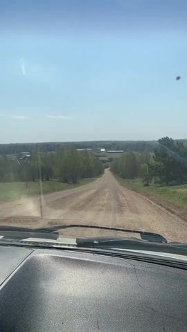 The video shows a car's perspective driving on a dirt road. The road is slightly uphill and leads to a distant view of buildings and trees. The sky is light blue with some clouds. The car's dashboard is visible in the foreground.