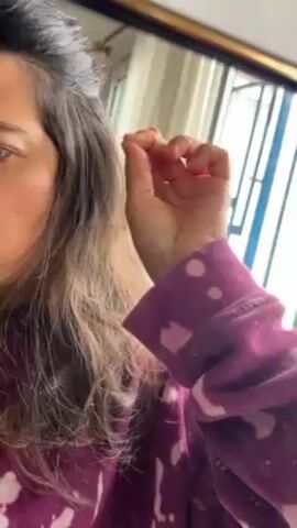 Here's a description of the video:
A woman stands indoors near a window, facing slightly towards a mirror. Her upper body is visible. She has long, dark brown hair that is partially gray at the roots. She is wearing a purple sweatshirt with a white splattered pattern. With her right hand, she is pinching her hair, close to her face, appearing to examine or adjust it. The window behind her has white curtains and a barred exterior.