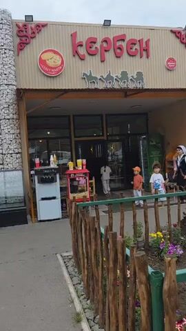 The video shows the exterior of two adjacent businesses. The business on the right has a sign that reads "Кербен" (Kerben) in red letters, with an illustration of camels above the entrance. There is a popcorn machine and an ice cream machine outside the entrance. Two children, one wearing an orange cap and the other a blue shirt, are standing near the entrance. A woman in traditional clothing is also visible. The business on the left has a sign with red letters that reads "Нурбўғ" (Nurbugh) above a smaller sign that reads "КАЙТАМА" (Kaitama). This building is covered in a stone-like material. An elderly woman with a cane is sitting outside the entrance to this building. In the background, there are some outdoor cooking setups with large pots. A wooden fence with a flowerbed in front of it separates the businesses from the viewer. The ground is paved, and the sky appears overcast.