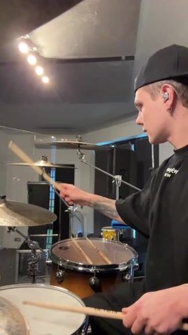 A medium shot shows a young man playing the drums in what appears to be a music studio. The drummer is wearing a black long-sleeved shirt and a backwards black baseball cap, and has a visible earbud and tattoos on his left arm. He is facing the right side of the frame, striking the cymbal with his right drumstick and smiling at the camera. The drum set, primarily consisting of a large snare drum, cymbals, and other drums, is situated in front of him. Behind the drummer is a dimly lit room with soundproofing material on the walls and a bright light overhead. A transparent drum screen is partly visible around the kit.