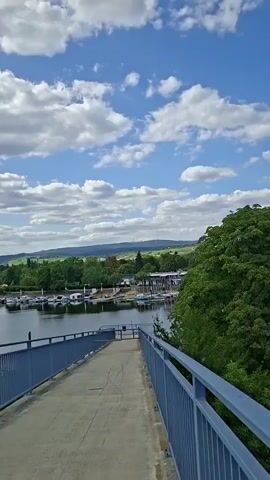 The video opens with a view of a walkway with a blue railing leading towards a body of water. Several boats are docked in the water, and lush green trees and rolling hills form the background. The sky is a bright blue with scattered white clouds. The camera pans slightly to the right, revealing more of the trees and the boats. The scene is peaceful and scenic, suggesting a marina or waterfront area on a pleasant day.