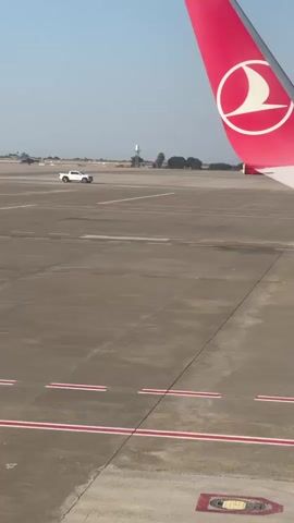 A white pickup truck drives across an airport tarmac. In the foreground, part of an airplane wing with the Turkish Airlines logo is visible. The sky is clear and blue, and the ground is paved concrete.