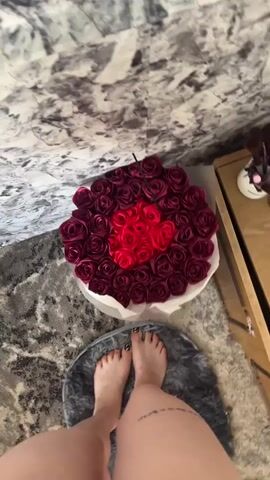 The video shows a top-down view of a person's feet standing on a round, grey rug. In front of the feet is a large bouquet of artificial roses. The roses are predominantly a deep burgundy color, arranged in a circular pattern. In the center of the bouquet, there are red roses arranged in a heart shape. The bouquet is in a white box. Behind the bouquet is a wall with a marble-like pattern. To the right of the bouquet, a wooden piece of furniture is visible, with a small white object on it.