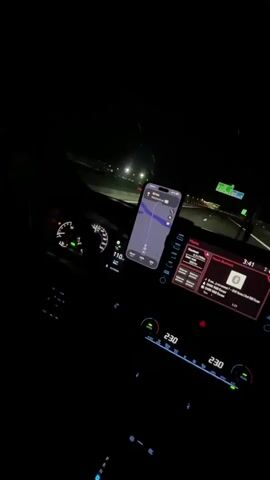 The video shows the interior of a car at night, as seen from the passenger's perspective. The dashboard is lit up, displaying the car's speed at 110 km/h and the time as 3:41 AM. A smartphone is mounted on the dashboard, displaying a navigation app with a route highlighted. The infotainment screen shows a music player interface. The road ahead is visible through the windshield, with streetlights illuminating the highway. The driver's side of the car is not fully visible, but the steering wheel and part of the driver's arm are discernible. The overall atmosphere is dark, with the car's interior lights and the illuminated screens being the primary light sources.