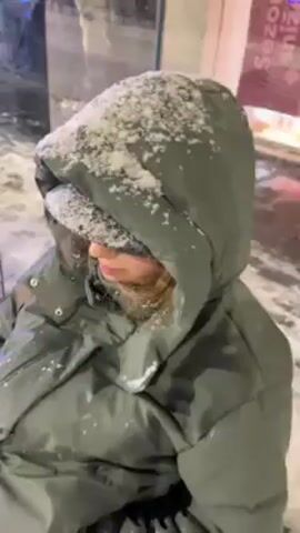 A young woman wearing a large, hooded winter coat and a bucket hat covered in snow looks at the camera and smiles slightly. She is likely indoors, as there is a reflection of indoor lighting behind her. The rest of her face is visible, though red from the cold. Her hair is light brown and also covered in snow.
