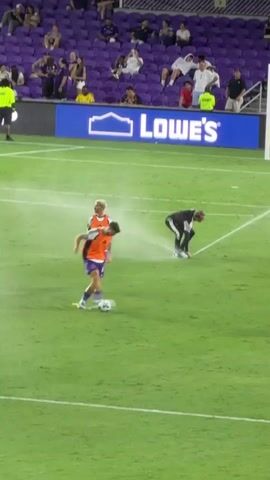 Two soccer players in uniform are on a green field. One player is wearing an orange vest over a purple uniform and white shoes, with the number 7 visible on the vest. The other player is wearing a black uniform with white accents and black shoes. The player in the black uniform appears to be spraying water from a hose onto the field near the player in the orange vest. In the background, there are spectators in purple seats and a large blue advertisement banner that says "LOWE'S". There are white lines marking the soccer field. A person wearing a bright yellow vest stands on the far left.