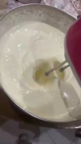 Here's a detailed description of the video:

The video shows someone using an electric hand mixer to whisk a white, creamy substance in a stainless steel mixing bowl. The mixer is predominantly pink. The bowl sits on a wooden surface, and in the background, there are glimpses of other objects, including what appears to be a box of whipped cream, a glass, and a decorative tablecloth. The cream is glossy, and the mixer is actively blending it, creating ripples in the mixture.