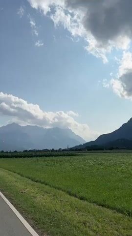 The video is a landscape shot featuring a vast green field under a bright, partially cloudy sky. In the distance, a range of majestic mountains forms the backdrop, their peaks partially obscured by clouds. A narrow paved road is visible in the foreground on the left side of the frame, bordered by a strip of grass. The overall impression is one of a serene rural setting.