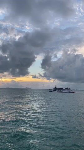 The video shows a landscape view of a calm, blue ocean with a ferry moving across the water. The sky above is filled with a mix of gray and white clouds, with a sliver of sunset visible through the cloud cover. The horizon features a faint outline of land, possibly islands, in the distance. The ferry is white and black, appearing to carry passengers. The overall scene has a serene and tranquil feel.