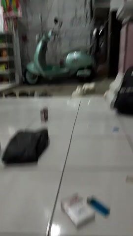 The video captures a ground-level view of a tiled floor in what appears to be a sheltered area, possibly a garage or an outdoor patio, in Panjalin Kidul, Indonesia. A light blue Vespa scooter is parked in the background, leaning on its kickstand. To the left of the scooter, a white shelving unit with various items is visible, suggesting a storage or retail space.

On the tiled floor, several objects are scattered. A black plastic bag lies to the left of the frame, with a bottle of water placed next to it. Further to the right, a pack of "CLAS" cigarettes and a blue lighter are prominently displayed, indicating a moment of pause or relaxation. A small, brown box, possibly a carton of cigarettes or a similar item, is positioned between the black bag and the scooter. A pair of white sneakers is placed near the scooter, along with several pairs of black sandals arranged in a line.

The scene is likely set during daylight hours, although the lighting is somewhat dim, suggesting it might be overcast or an interior space. The overall impression is one of a casual, everyday setting, with personal belongings laid out as if in preparation for or after an activity. There are no people clearly visible in the frame, adding to the sense of quiet observation. The presence of the scooter and the general arrangement of items suggest a residential or mixed-use area. The visible text on the cigarette pack is "CLAS".