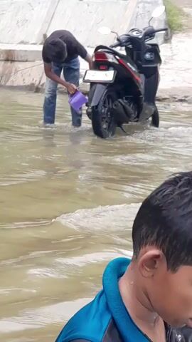 A person in a dark shirt and blue jeans is standing in shallow, murky water, scooping water with a purple ladle. They are bent over and appear to be pouring the water onto the back tire of a black scooter parked in the water. The scooter has a license plate that is partially visible. Another person's head and shoulders are visible in the foreground, out of focus, wearing a blue shirt with a dark collar. The background shows a concrete structure and some greenery. The water is rippling and appears to be flowing.