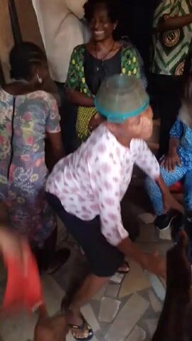The video captures a lively social gathering indoors. A woman in a white shirt with pink patterns and a green brimmed hat dances enthusiastically in the center. She’s surrounded by other people, some sitting, others standing, observing and engaging with the activity. Many wear colorful, patterned clothing typical of traditional or cultural attire. The environment appears to be a home or community space. The focus is clearly on the woman dancing and the communal celebration taking place.