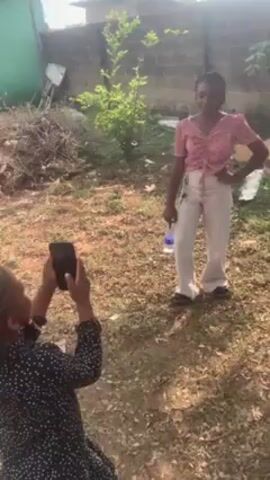 A young girl in a black dress with white dots is filming a teenage girl with a phone. The teenage girl is wearing a light pink shirt and white pants. She is holding a water bottle in one hand and posing with her other hand on her head. They are standing in a grassy area with some brush and a brick wall in the background.