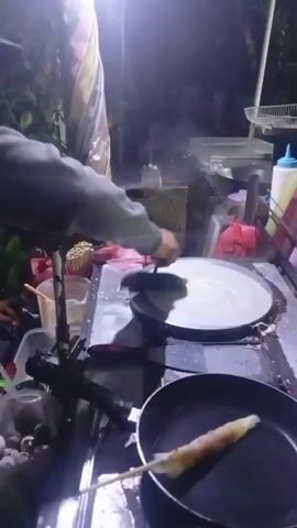 A man is preparing food at a street stall at night. He is wearing a dark grey hoodie and a black baseball cap. He is using a spatula to spread batter on a flat cooking surface, possibly a crepe maker. In the foreground, there is a frying pan with a rolled-up food item on a skewer. Several containers of ingredients are visible, including a container of what appear to be quail eggs. The scene is lit by artificial lights, creating a dimly lit atmosphere with shadows. Lush green foliage is visible in the background.