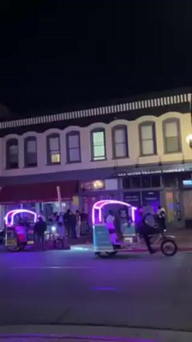 The video shows a street corner scene at night. People walk across the intersection, and cars drive through. A neon-lit bicycle taxi stands at the corner. The buildings are historic, with arched windows and illuminated facades. There are streetlights, traffic signals, and numerous pedestrians. A police car with flashing lights is visible further down the street. The overall atmosphere is lively and urban.