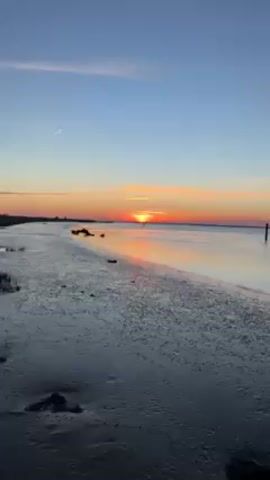 A sunrise over water is seen. The sky is a blend of blues, oranges, reds, and yellows.  The sun is low on the horizon, appearing as a bright orange disk with dark lines of clouds above it. Its reflection shimmers on the surface of the calm water.  Dark posts stick out of the water at intervals.  The edge of a wet, sandy or muddy beach is visible in the foreground, dotted with small debris. The camera slowly pans upward, emphasizing the sky and the vibrant colors of the sunrise.