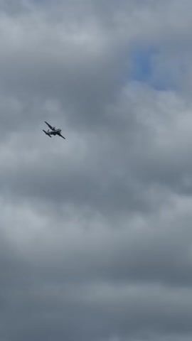 A dual-engine propeller airplane flies through a cloudy sky. The plane is white with dark accents and is seen from below and slightly to the side. The clouds are gray and cover most of the frame, with occasional patches of blue sky visible. The plane is the only object in the frame and appears to be in mid-flight, at a moderate altitude.