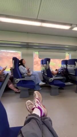 The interior of a train car is shown from the perspective of a seated passenger with their feet up on the seat. The train car has gray walls, a white ceiling with small holes, and blue seats. Three windows show green foliage passing by outside. Four other passengers are visible: one older woman on the left, a young woman looking at her phone, and a man in a white shirt and jeans. All appear to be using electronic devices. The seated passenger has on grey pants, white socks, and worn athletic shoes.