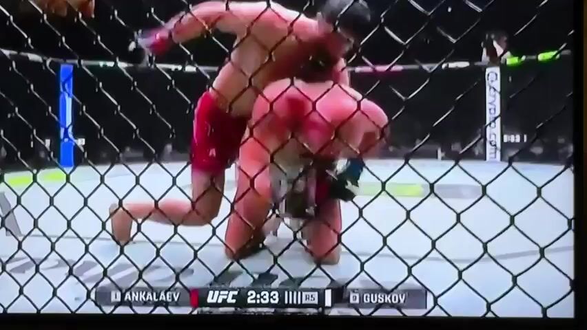This is a video of an MMA fight taking place inside a cage. Two male fighters are on the ground, entangled. One fighter, wearing red shorts, is in a dominant position on top of the other fighter. The fighter on the bottom is wearing black shorts. The fight is being broadcast on a television screen, with a UFC logo and fighter names, Magomed Ankalaev and Ion Cutelaba, visible at the bottom of the screen, along with a timer indicating 2:28 remaining in the round. The background appears to be a dimly lit arena with spectators.