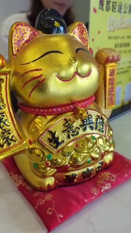 A gold maneki-neko, or lucky cat figurine, is displayed on a red cushion with a floral pattern. The cat is facing forward with its right paw raised and its eyes closed in a smile. The figurine is adorned with intricate gold designs and Chinese characters. In the background, a yellow sign with Chinese text and the word "Welcome" is visible. The lighting is soft and appears to be indoors. The video appears to be a still shot or a very slow pan over the figurine.