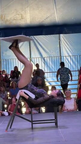 A woman in a sparkling leotard lies on her back on a black chair, performing an acrobatic balancing act. Her legs are extended upwards, and she is balancing two white plates, one on each foot. The performance takes place on a stage, with an audience seated in the background. The stage is covered with a blue tarp, and a metal railing separates the stage from the audience. The audience members are diverse in age and are watching the performance with interest.