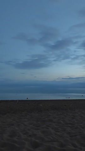 A low-angle, vertical, wide shot shows a dark, sandy beach in the foreground. The sand is rippled and textured. In the middle ground, a calm sea stretches horizontally across the frame. Small, round, red buoys are visible floating in the water, one in the left half of the frame and two more further to the right. The sea meets a hazy horizon line beneath a vast, blue-grey sky filled with scattered clouds. The overall lighting suggests dusk or early dawn, with a soft, diffused light. The scene is peaceful and empty.