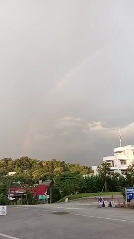 A short video shows a rainbow against a cloudy sky. In the foreground, there is a paved area with a few signs and a white building with a flagpole. The building appears to be a school or hospital. There are trees and houses in the background. The video is taken from a slightly elevated position, looking across a road and towards the building and rainbow.